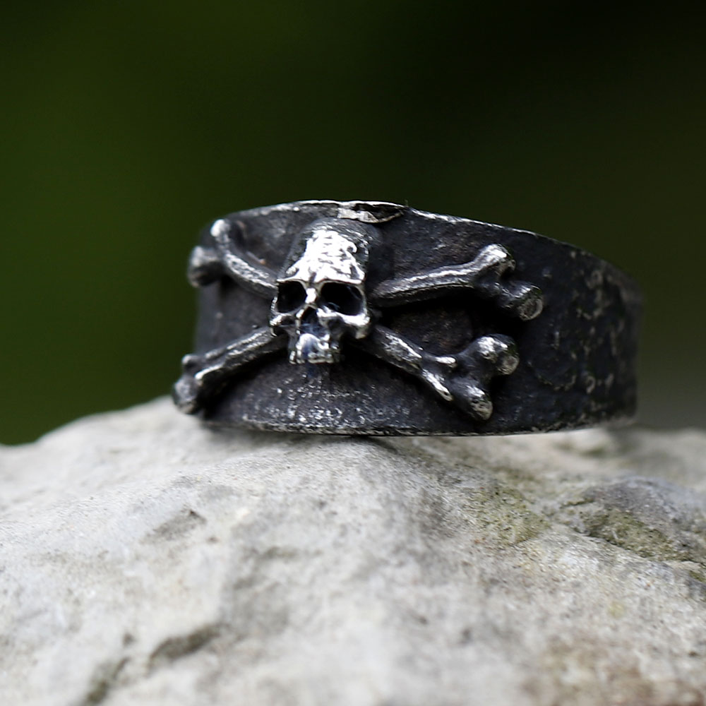Frontal close-up of the men's skull and crossbones ring, showcasing its wide band and aged finish on wood.