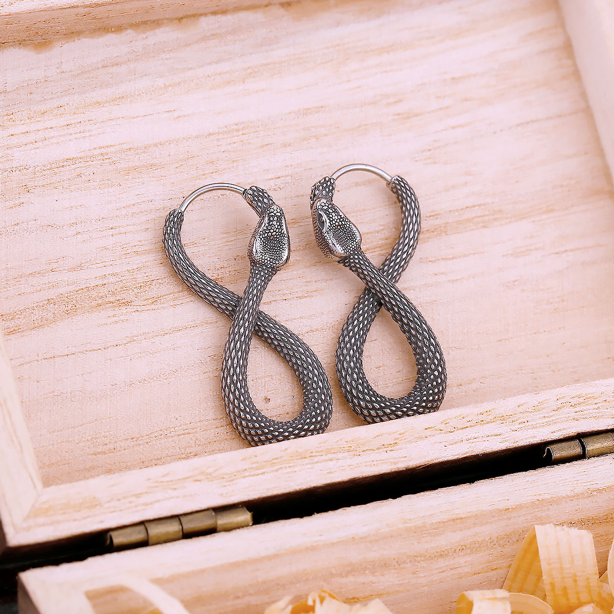 A pair of stainless steel snake earrings, sculpted into a figure-eight, resting in a light wooden box.